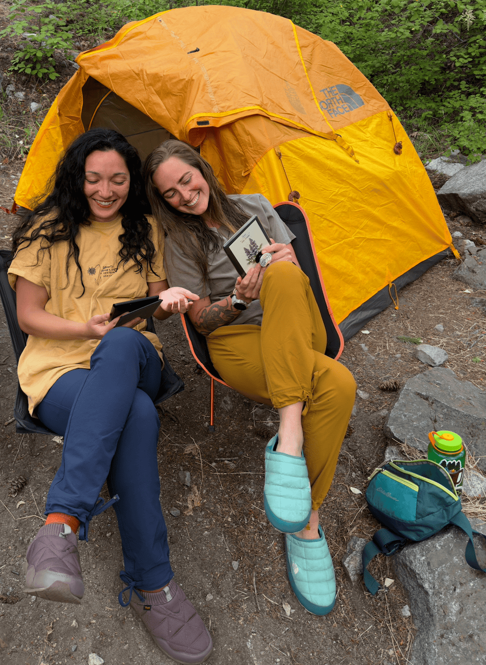 Two friends reading and laughing while reading their Kindles on a backpacking trip. These Coalatree Trailhead pants are a perfect item if you're deciding what to wear in Seattle in the fall.